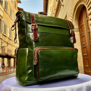 May include: A green leather backpack with brown leather straps and gold-coloured buckles. The backpack has a front pocket with a brown zip and a small logo. The text "ITALYLEATHERMAN" is visible at the top.
