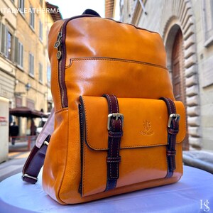 Op de afbeelding: Een oranje leren rugzak met donkerbruine details, waaronder riemen en gespen. De tas heeft een voorvak met twee gespsluitingen en een handvat aan de bovenkant. De tekst "ITALYLEATHERMA" is bovenaan zichtbaar.