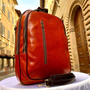 Peut inclure: Un sac à dos en cuir marron-rouge avec des fermetures éclair et des bordures marron foncé. Le sac a une poignée supérieure et une bandoulière amovible. Le fond montre un bâtiment de style européen. Le texte "ITALY LEATHER" est visible en haut.
