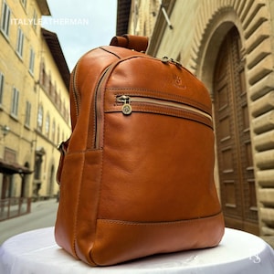 Puede incluir: Una mochila de cuero marrón con cremalleras y herrajes dorados. La mochila tiene un bolsillo frontal con cremallera y un asa superior. La marca "ITALY LEATHERMAN" es visible en la esquina superior izquierda.