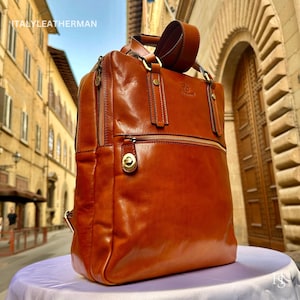 May include: A brown leather backpack with a top handle and adjustable straps. The bag has a front zipper pocket and gold-colored hardware. The brand name "ITALY LEATHERMAN" is visible at the top.