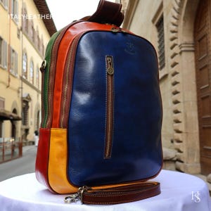 May include: A multi-coloured leather backpack featuring a large blue front panel with a vertical zipped pocket, a yellow base, a red side panel, a green side panel, and brown trim. The bag includes brass-toned hardware and a detachable brown strap.