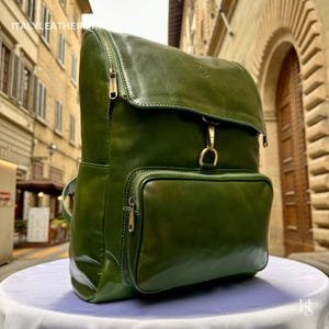 May include: A green leather backpack with a flap closure and a front zipped pocket. The backpack has gold-coloured hardware and a top handle. The bag is on a white surface.