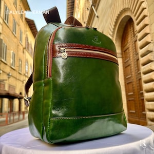May include: Green leather backpack with brown accents and a gold zipper. The backpack has a top handle and adjustable straps. The text "ITALYLEATHERMAN" is visible at the top. The backpack is sitting on a white surface.