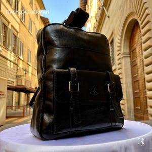 May include: Black leather backpack featuring a front pocket with two buckle-fastened straps. The bag has a top handle and adjustable shoulder straps. The text "ITALY LEATHERMAN" is visible at the top of the bag.