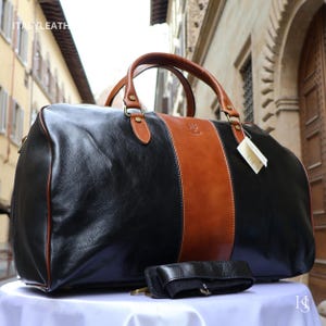 May include: A black and tan leather duffel bag featuring two top handles, a central vertical tan stripe, and a detachable black shoulder strap. The bag has a smooth finish, gold-tone hardware, and a white tag attached to one of the handle rings.