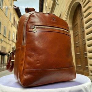 Op de afbeelding: Een bruine leren rugzak met een voorvak met ritssluiting en goudkleurige hardware. De rugzak heeft verstelbare bandjes en een handvat aan de bovenkant. De merknaam "ITALY LEATHERMAN" is zichtbaar bovenaan. De tas staat op een wit oppervlak.