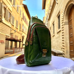 May include: A green leather sling bag with brown trim and strap. The bag features a front pocket with a clasp and two zipped compartments. The text "ITALY LEATHERMAN" is visible at the top left.