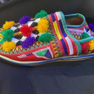 Könnte beinhalten: Ein farbenfroher, handgefertigter Slipper mit roter Basis, verziert mit lebendiger Stickerei und Pompons. Das Design zeigt ein Schachbrettmuster und eine Mischung aus grünen, gelben, lila und roten Akzenten. Die Sohle ist flach und der Schuh hat eine runde Spitze.