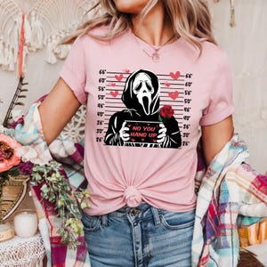May include: Pink t-shirt featuring a black and white graphic of a Scream mask holding a sign that reads "NO YOU HANG UP." The design includes red hearts and a red rose. The shirt has short sleeves and a tied front.