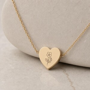 Puede incluir: Un collar con colgante en forma de coraz&oacute;n de tono dorado que incluye una cadena fina y delicada. La superficie del coraz&oacute;n presenta un grabado de l&iacute;neas finas que muestra una flor sencilla con cinco p&eacute;talos y hojas.