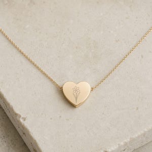 May include: A gold-toned necklace featuring a heart-shaped pendant with a delicate, engraved line drawing of a daffodil flower. The pendant is centered on a thin, matching gold-toned chain that connects to both sides of the heart-shaped charm.