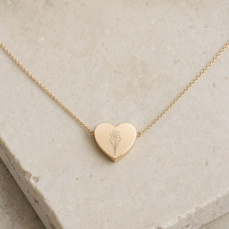 May include: A gold-toned necklace featuring a heart-shaped pendant with a delicate, engraved line drawing of a daffodil flower. The pendant is centered on a thin, matching gold-toned chain that connects to both sides of the heart-shaped charm.
