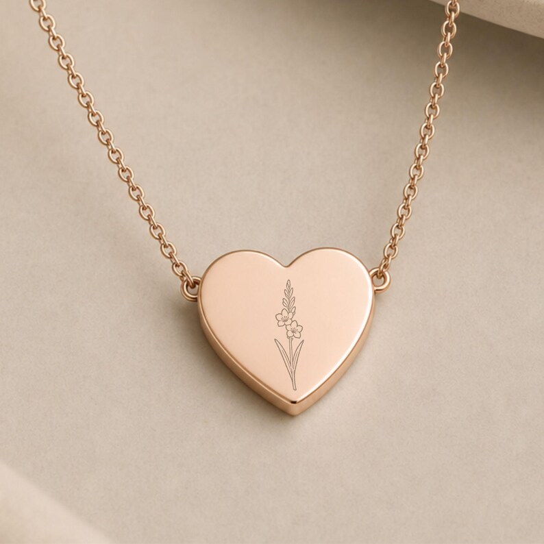 May include: A rose gold heart-shaped pendant necklace featuring a delicate, thin-line engraving of a single flower stem with small blossoms centered on the front surface, suspended from a matching fine link chain.