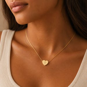 Woman wearing a gold carnation birth flower heart necklace at her collarbone over a cream ribbed tank top
