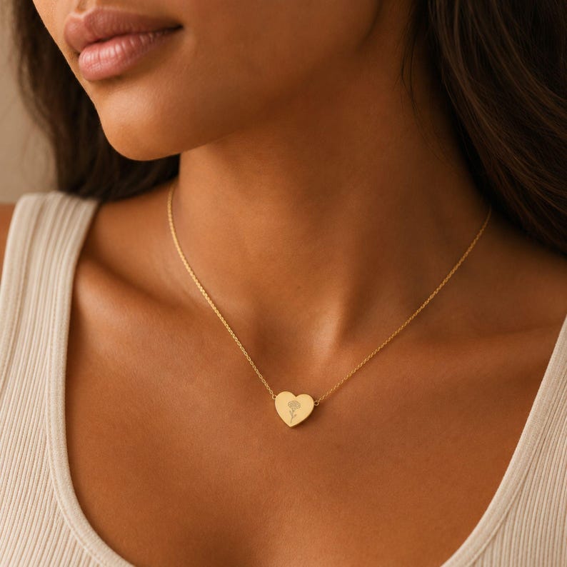 Woman wearing a gold carnation birth flower heart necklace at her collarbone over a cream ribbed tank top
