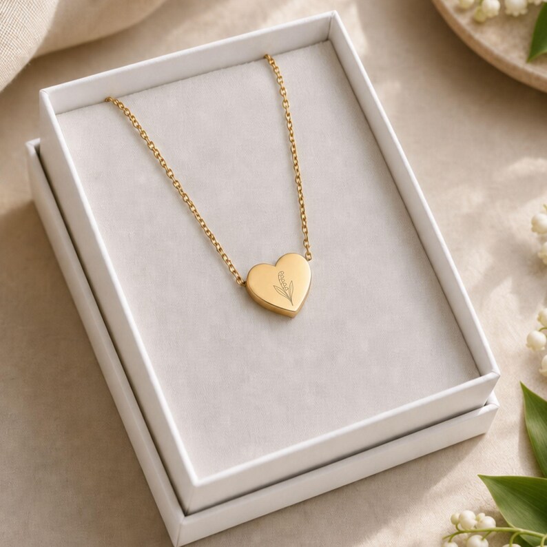 Pu&ograve; includere: Una collana color oro con un ciondolo a forma di cuore caratterizzato da un delicato disegno floreale inciso. La collana &egrave; presentata all'interno di una scatola regalo rettangolare bianca con rivestimento interno grigio, che mostra la catena.