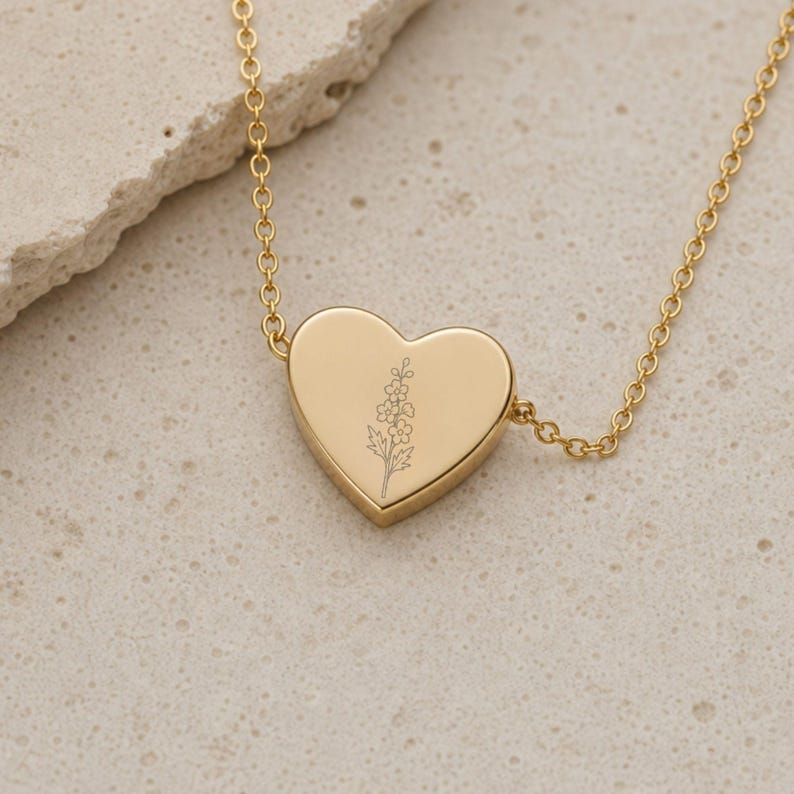 Puede incluir: Un collar de tono dorado con un colgante en forma de coraz&oacute;n que presenta un delicado dibujo lineal grabado de una flor silvestre. El colgante est&aacute; centrado en una cadena fina y pulida de tono dorado, creando una pieza de joyer&iacute;a sencilla.