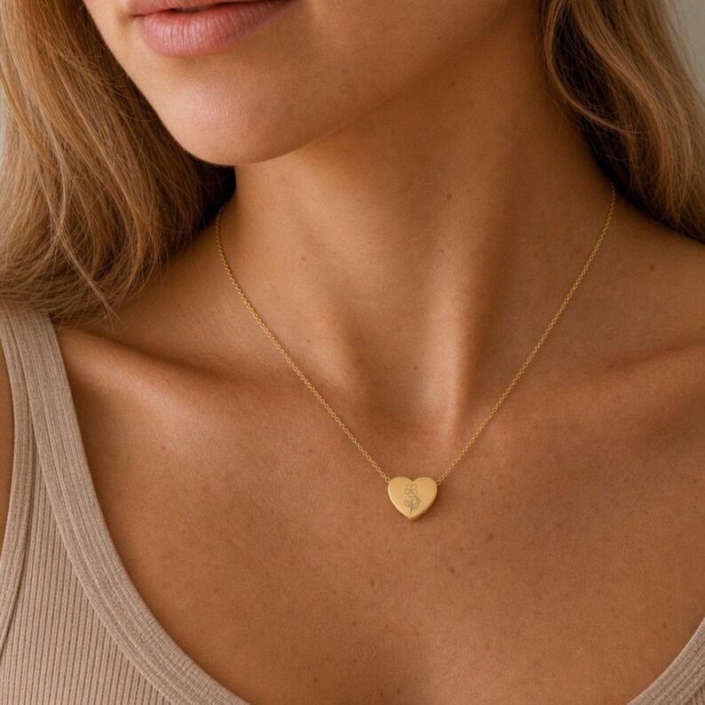 Puede incluir: Un collar de tono dorado que presenta un peque&ntilde;o colgante en forma de coraz&oacute;n suspendido de una cadena delicada. El centro del colgante de coraz&oacute;n est&aacute; grabado con un dibujo sencillo de l&iacute;neas finas de una flor con tallo y dos hojas.