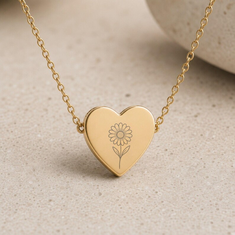 May include: A gold-toned necklace featuring a heart-shaped pendant with a centered, engraved line drawing of a daisy flower. The pendant is attached to a delicate gold-toned chain that extends from both sides of the heart, creating a simple, polished look.