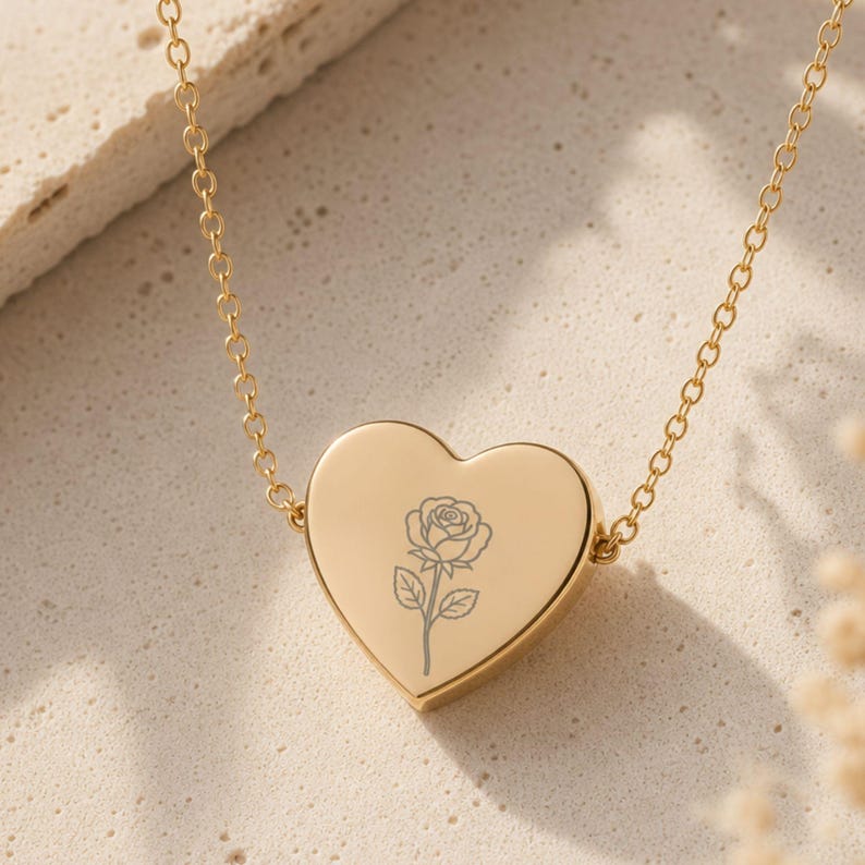 May include: A gold-toned heart-shaped pendant necklace featuring a delicate, engraved line drawing of a single rose centered on the front surface. The pendant is suspended from a thin, matching gold-toned cable chain with circular links.