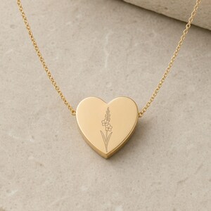 May include: A gold-toned heart-shaped pendant necklace featuring a delicate, fine-line engraving of a single floral stem. The smooth, polished metal heart is suspended from a thin, matching gold-toned chain, creating a simple and elegant piece of jewelry.