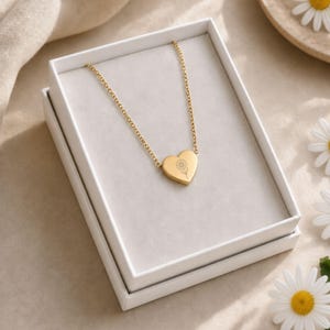 May include: A gold-toned necklace featuring a heart-shaped pendant with an engraved sunflower design. The pendant is suspended from a delicate gold-toned chain, and the entire piece is presented inside a white rectangular gift box.