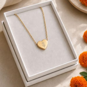 May include: A gold-toned necklace featuring a heart-shaped pendant with a delicate line-art flower engraving. The pendant is suspended from a thin gold chain and is presented inside a white rectangular gift box with a soft grey interior lining.
