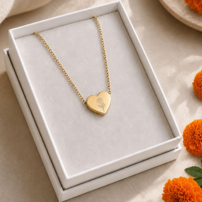 May include: A gold-toned necklace featuring a heart-shaped pendant with a delicate line-art flower engraving. The pendant is suspended from a thin gold chain and is presented inside a white rectangular gift box with a soft grey interior lining.