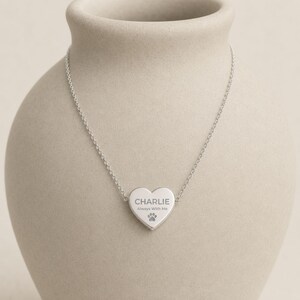 May include: A silver-toned necklace featuring a heart-shaped pendant. The pendant is engraved with the name CHARLIE, the phrase Always With Me, and a small paw print icon. The pendant is attached to a delicate, thin silver-toned chain.