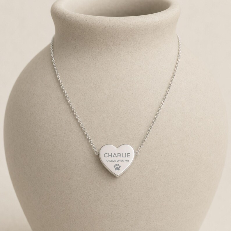 May include: A silver-toned necklace featuring a heart-shaped pendant. The pendant is engraved with the name CHARLIE, the phrase Always With Me, and a small paw print icon. The pendant is attached to a delicate, thin silver-toned chain.