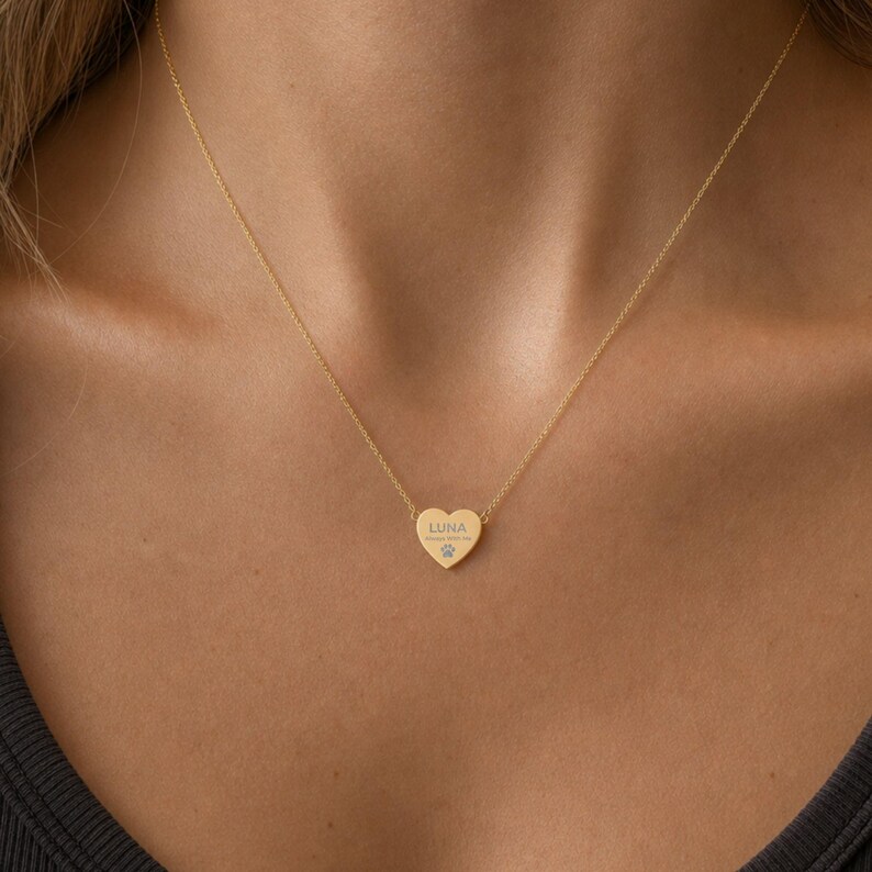 May include: A gold-toned heart-shaped pendant necklace featuring the engraved name LUNA, the text Always With Me, and a small paw print icon. The pendant is suspended from a delicate gold-toned chain that rests against the collarbone area.