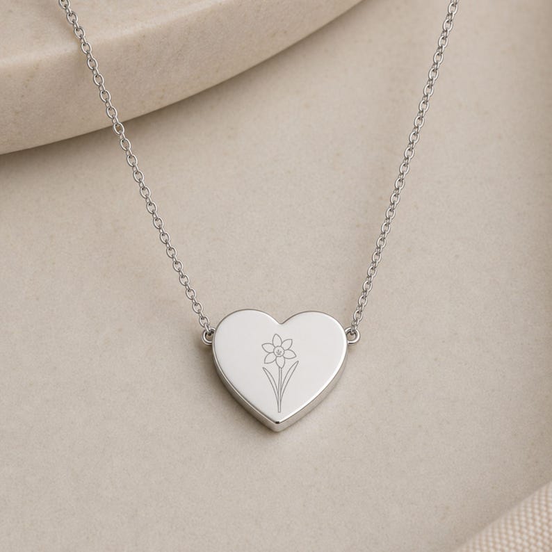 May include: A silver-toned heart-shaped pendant necklace featuring a delicate, fine-line engraving of a single daffodil flower centered on the front surface. The pendant is suspended from a thin, matching silver-toned metal chain link necklace.