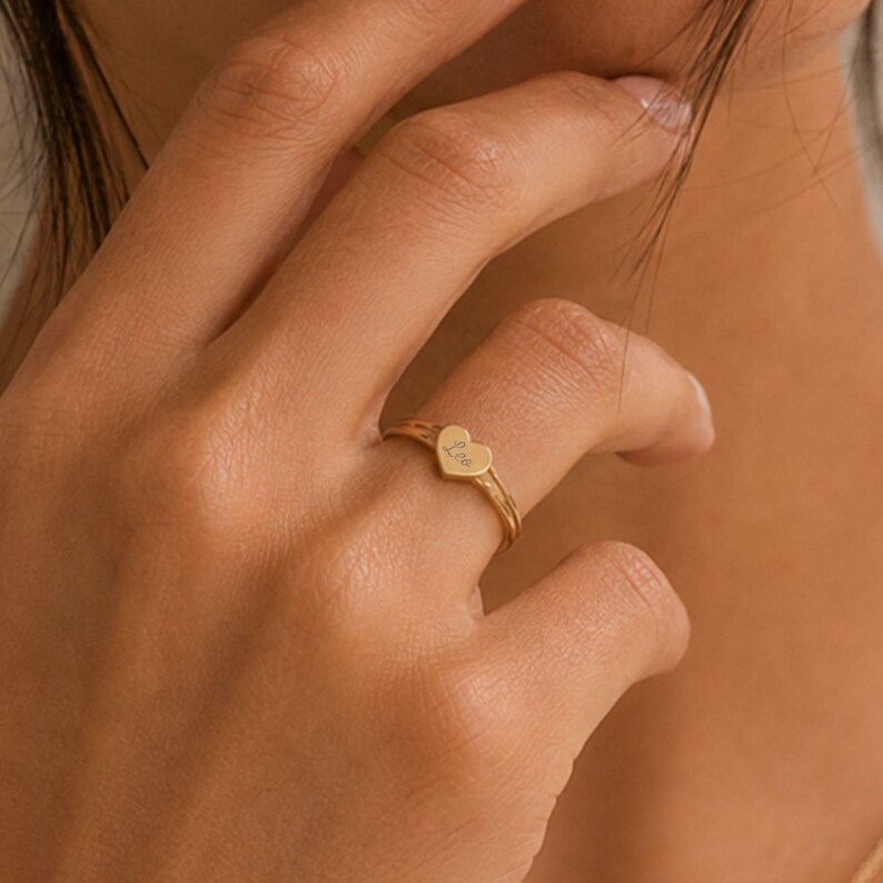 Puede incluir: Un anillo de tono dorado que presenta un peque&ntilde;o dije plano en forma de coraz&oacute;n centrado en una banda delgada y texturizada. El dije de coraz&oacute;n est&aacute; grabado con el nombre Leo en letra cursiva. El anillo tiene un acabado pulido y fino.