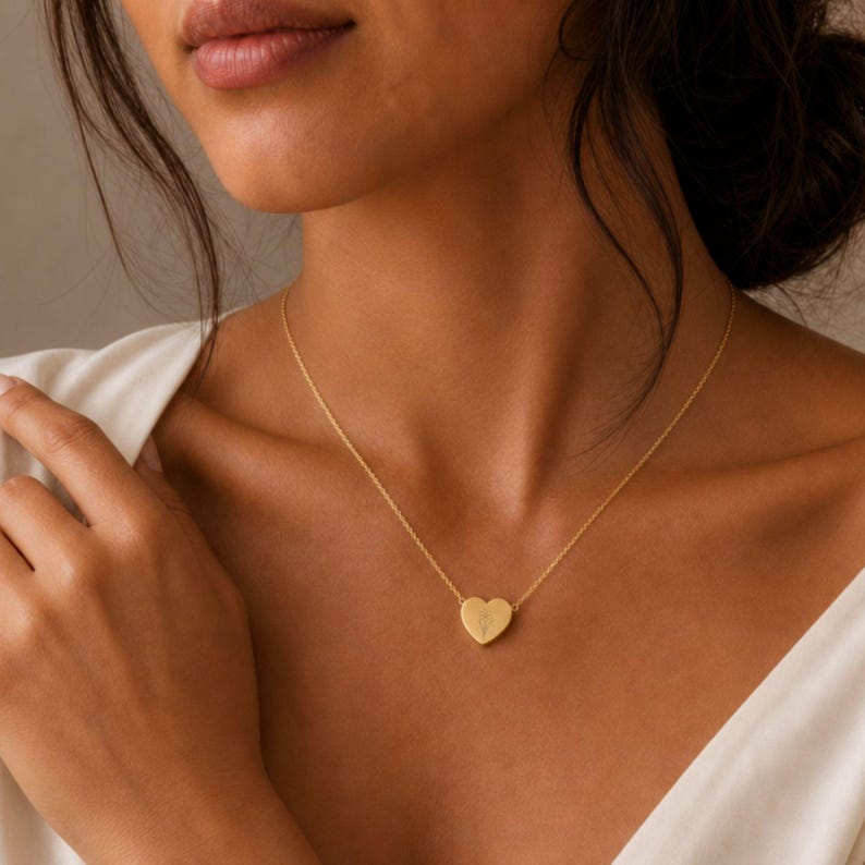 May include: A gold-toned necklace featuring a delicate heart-shaped pendant. The pendant is suspended from a thin, matching gold-toned chain. A small, subtle botanical engraving is visible on the center of the heart-shaped charm, which rests against the collarbone.