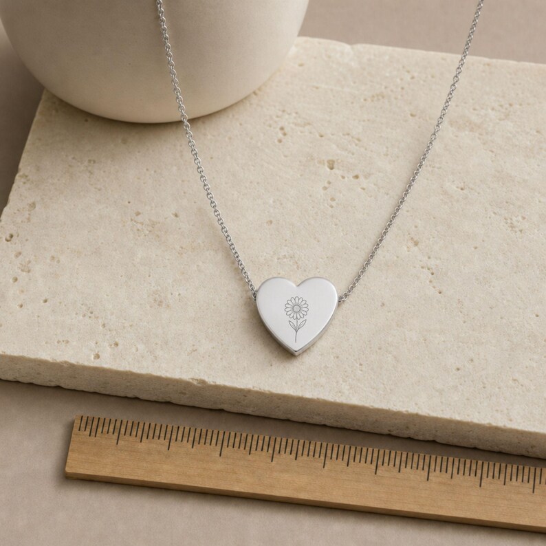 May include: A silver-toned heart-shaped pendant necklace featuring a delicate engraved daisy flower design. The pendant is attached to a thin metal chain. A wooden ruler is positioned below the necklace to show the scale of the jewelry piece.