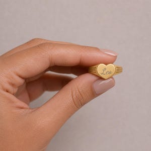 Puede incluir: Un anillo de tono dorado con una cara en forma de coraz&oacute;n que tiene el nombre Leo grabado en letra cursiva. La banda del anillo muestra una serie de l&iacute;neas verticales grabadas a ambos lados del coraz&oacute;n, creando un detalle texturizado en el metal.