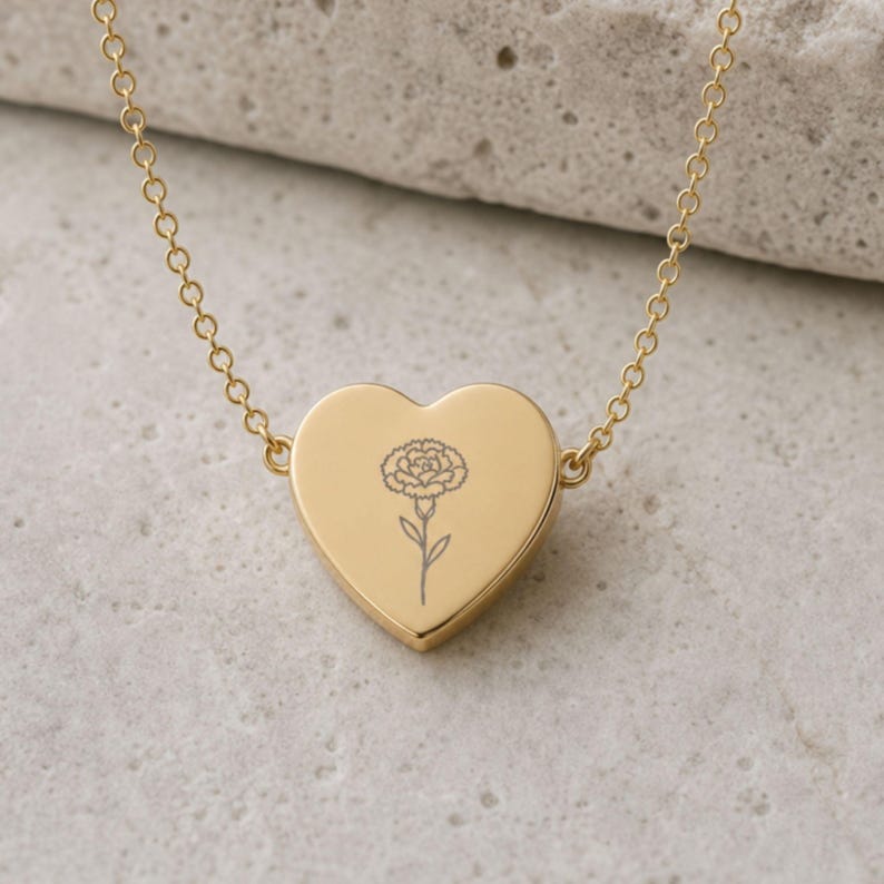 Gold carnation necklace with a dainty heart pendant engraving of the January birth flower, on a stone surface