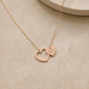 May include: A rose gold-toned necklace featuring a delicate chain and two heart-shaped pendants. One pendant is an open heart outline, while the smaller, solid heart pendant is engraved with the letters J & C in a simple, clean font.