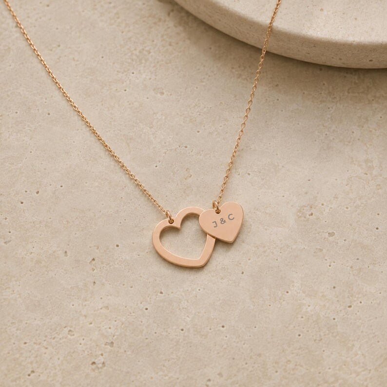 May include: A rose gold-toned necklace featuring a delicate chain and two heart-shaped pendants. One pendant is an open heart outline, while the smaller, solid heart pendant is engraved with the letters J & C in a simple, clean font.