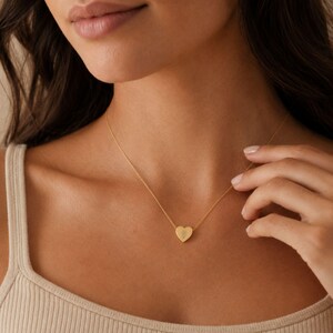 May include: A gold-toned necklace featuring a delicate chain and a small, smooth heart-shaped pendant. The pendant is centered on the collarbone and displays a subtle, engraved floral design on its surface, reflecting a soft metallic sheen.