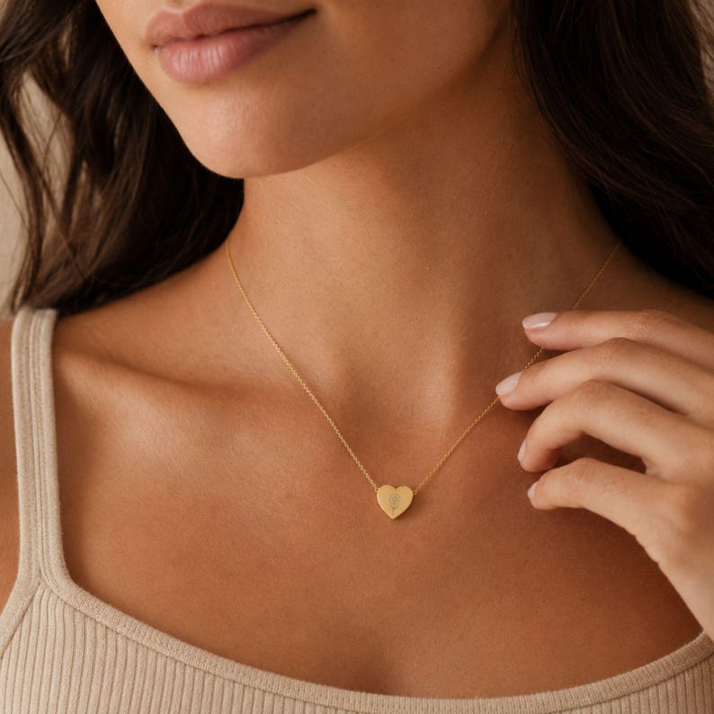 May include: A gold-toned necklace featuring a delicate chain and a small, smooth heart-shaped pendant. The pendant is centered on the collarbone and displays a subtle, engraved floral design on its surface, reflecting a soft metallic sheen.