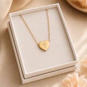 Gold carnation heart pendant necklace in a white gift box beside fresh carnations, a January birthday gift