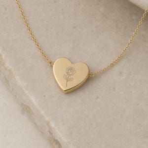 May include: A gold-toned necklace featuring a heart-shaped pendant with a delicate, engraved line drawing of a single rose centered on its surface. The pendant is attached to a thin, matching gold-toned chain that extends to both sides.