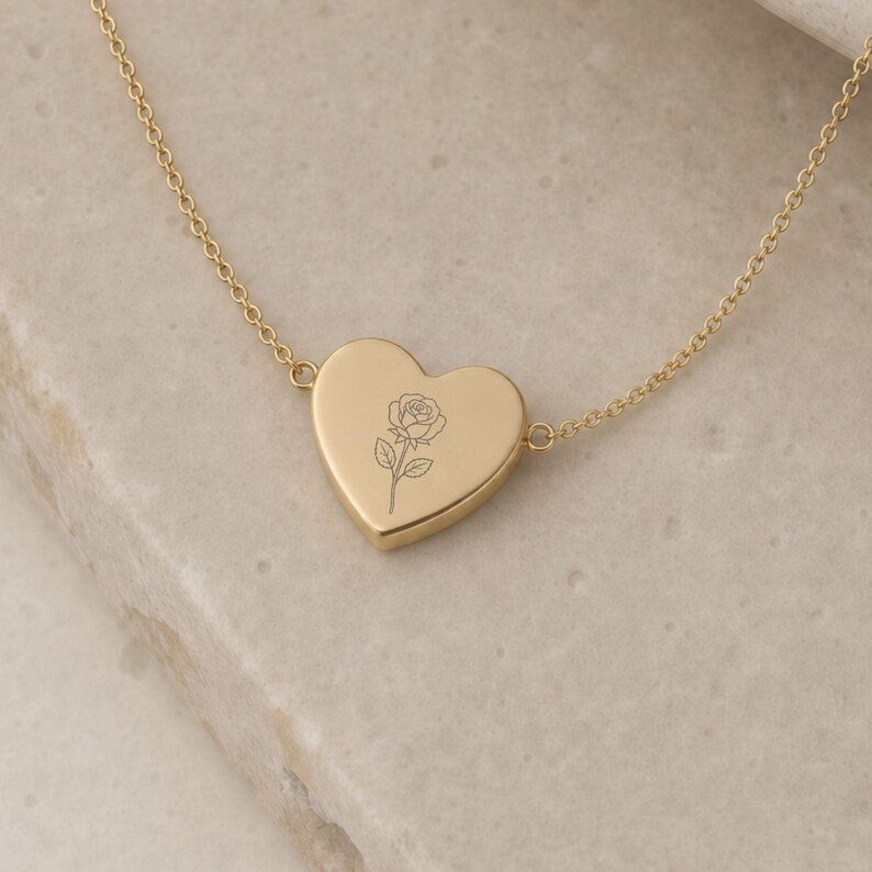 May include: A gold-toned necklace featuring a heart-shaped pendant with a delicate, engraved line drawing of a single rose centered on its surface. The pendant is attached to a thin, matching gold-toned chain that extends to both sides.
