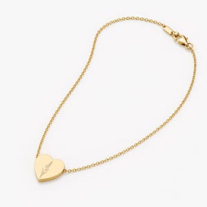 May include: A gold-toned chain bracelet featuring a heart-shaped charm. The charm is engraved with a delicate, thin-lined floral illustration. The bracelet includes a standard lobster claw clasp mechanism at the end of the fine metal chain links.
