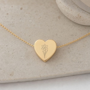 May include: A gold-toned heart-shaped pendant necklace featuring a delicate, fine-line engraving of a single daffodil flower centered on the front surface. The pendant is suspended from a thin, matching gold-toned metal chain link necklace.