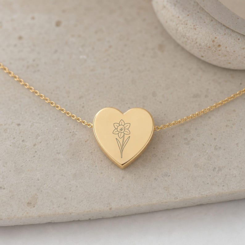 May include: A gold-toned heart-shaped pendant necklace featuring a delicate, fine-line engraving of a single daffodil flower centered on the front surface. The pendant is suspended from a thin, matching gold-toned metal chain link necklace.