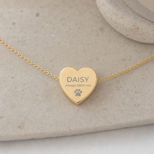 May include: A gold-toned heart-shaped pendant necklace featuring the engraved text DAISY, the phrase Always With Me, and a small paw print icon. The pendant is attached to a delicate gold-toned chain that extends horizontally across the frame.