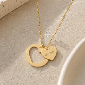 May include: A gold-toned necklace featuring a delicate chain and two heart-shaped pendants. One pendant is a solid gold heart outline, while the smaller, solid gold heart charm is engraved with the name Lindsey in a simple, dark font.